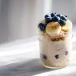 Glass jar of toddler overnight oats no added sugar, topped with banana slices and blueberries, on a white tabletop bathed in soft morning light.