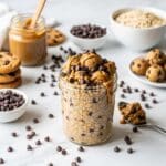 A professional, high-resolution horizontal food photography image for a WordPress blog post. The scene is shot from a 45-degree angle on a bright white marble countertop with playful, cheerful lighting. CENTER FOCUS: A thick glass jar overflowing with Cookie Dough Overnight Oats, showing golden cookie dough-colored oats studded with mini chocolate chips throughout, topped with edible cookie dough chunks and drizzled with creamy almond butter. Mini chocolate chips are scattered abundantly on top and around the jar. A spoon with cookie dough is positioned nearby. SUPPORTING ELEMENTS: Mini chocolate chips scattered generously across the marble, a small bowl filled with more chocolate chips, chocolate chip cookies stacked nearby, a jar of almond butter with a wooden spoon, rolled oats in a modern white bowl, and edible cookie dough balls on a small plate. COMPOSITION: Jar centered with slight offset to the left, leaving negative space for text overlay. LIGHTING: Bright, clean natural light makes the chocolate chips shine and creates an indulgent, dessert-like atmosphere. STYLE: Modern, fun food photography with a dessert-for-breakfast vibe. Color palette is golden cookie dough tones, dark chocolate browns, and bright white creating an indulgent yet wholesome look. IMAGE DIMENSIONS: 1200x630 pixels, optimized for WordPress featured image and social media sharing.