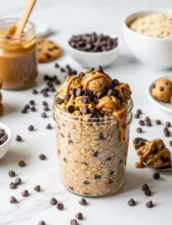 A professional, high-resolution horizontal food photography image for a WordPress blog post. The scene is shot from a 45-degree angle on a bright white marble countertop with playful, cheerful lighting. CENTER FOCUS: A thick glass jar overflowing with Cookie Dough Overnight Oats, showing golden cookie dough-colored oats studded with mini chocolate chips throughout, topped with edible cookie dough chunks and drizzled with creamy almond butter. Mini chocolate chips are scattered abundantly on top and around the jar. A spoon with cookie dough is positioned nearby. SUPPORTING ELEMENTS: Mini chocolate chips scattered generously across the marble, a small bowl filled with more chocolate chips, chocolate chip cookies stacked nearby, a jar of almond butter with a wooden spoon, rolled oats in a modern white bowl, and edible cookie dough balls on a small plate. COMPOSITION: Jar centered with slight offset to the left, leaving negative space for text overlay. LIGHTING: Bright, clean natural light makes the chocolate chips shine and creates an indulgent, dessert-like atmosphere. STYLE: Modern, fun food photography with a dessert-for-breakfast vibe. Color palette is golden cookie dough tones, dark chocolate browns, and bright white creating an indulgent yet wholesome look. IMAGE DIMENSIONS: 1200x630 pixels, optimized for WordPress featured image and social media sharing.